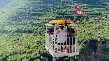 Tres personas viajan en una góndola abierta con techo naranja, suspendida por cables sobre un denso valle montañoso cubierto de vegetación. La bandera peruana ondea