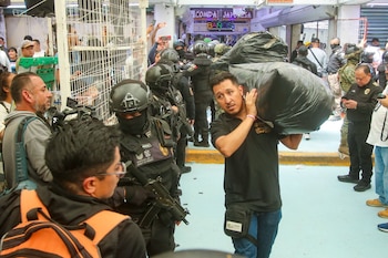 Hombres cargando grandes bolsas negras de mercancía incautada, rodeados de agentes de seguridad armados con uniformes negros y chalecos del Gobierno de la CDMX, en un pasillo comercial
