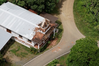 Vista aérea de una iglesia blanca con tejas dañadas en el techo, junto a una carretera asfaltada, un camino de tierra y varios árboles