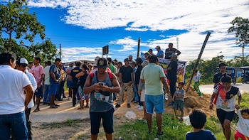 Multitud de personas, principalmente hombres, se congregan en un camino de tierra junto a una carretera, bajo un cielo azul con nubes blancas y vegetación