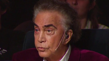 Primer plano del cantante José Luis Rodríguez con un micrófono de diadema y saco granate sobre un fondo oscuro