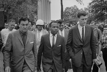 Fotografía en blanco y negro de James Meredith caminando en traje junto a varios hombres vestidos de traje en un entorno universitario