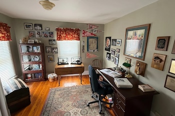 Un escritorio oscuro, silla de oficina, dos ventanas con cortinas rojas, una biblioteca, una mesa auxiliar, vitrinas y numerosas fotografías en la pared