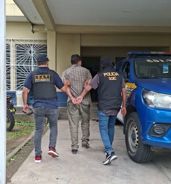 Dos agentes de policía llevan de los brazos a un hombre esposado caminando hacia una entrada junto a un vehículo policial azul