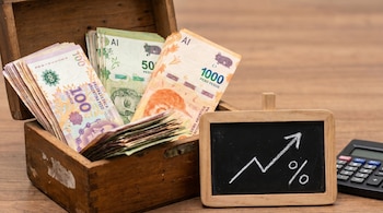Caja de madera con billetes de pesos argentinos (100, 50, 1000), junto a una pizarra que muestra una flecha ascendente y un porcentaje, y una calculadora.