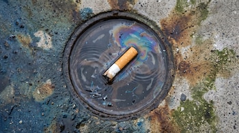 Vista cenital de una alcantarilla con un charco de agua, una colilla de cigarrillo flotando, manchas de aceite, siluetas de peces y microplásticos.