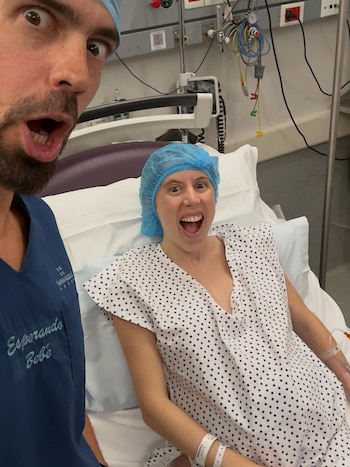 Una mujer con gorro y bata de hospital sonríe ampliamente en una cama. Un hombre con gorro y uniforme "Esperando Bebé" toma una selfie