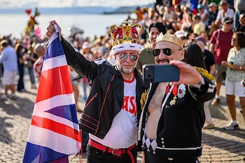 Dos hombres con coronas y gafas de sol posan para una foto; uno sostiene la bandera del Reino Unido frente a una multitud