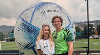 Guillermo Ochoa y Karla Mora posan sonriendo frente a un balón gigante blanco y azul con el logo de Adidas, en un campo de fútbol con una red.