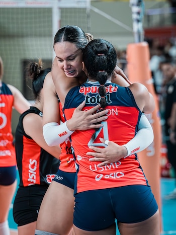Dos jugadoras de voleibol con camisetas rojas y azules se abrazan en una cancha. Una tiene lágrimas en el rostro, mientras otra jugadora es visible detrás