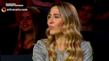 Plano medio de una mujer rubia con un micrófono de vincha y ropa brillante dentro de un estudio de televisión con público al fondo