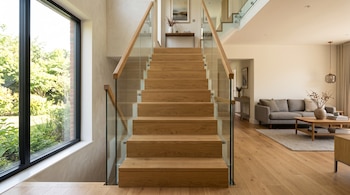 Escalera de madera con paneles de vidrio, un ventanal con vistas al jardín y una sala de estar con sofá y mesa al fondo.