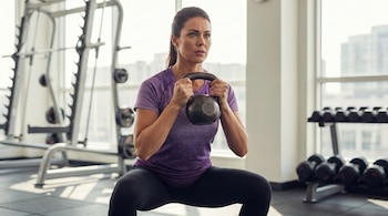 Imagen de una mujer de 45 años con remera violeta y pantalones oscuros haciendo sentadilla con una pesa rusa en un gimnasio iluminado por la luz natural.