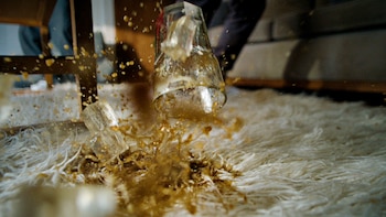 Primer plano de un vaso de bebida oscuro con hielo cayendo y derramándose sobre una alfombra blanca o crema de pelo largo, creando salpicaduras