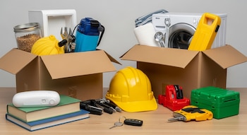 Dos cajas de cartón abiertas con electrodomésticos, enseres y herramientas. Sobre una mesa: libros, un casco de seguridad amarillo, herramientas y llaves de coche.