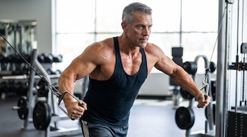 Un hombre con camiseta sin mangas utiliza una máquina de cables con agarres en un gimnasio rodeado de pesas.