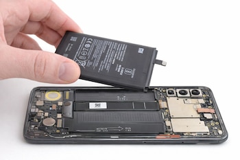 Primer plano de una mano extrayendo una batería negra de litio de un smartphone Xiaomi abierto, mostrando sus componentes internos sobre un fondo blanco.