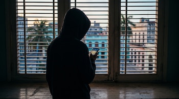 Silueta de espaldas de una persona con capucha que sostiene un teléfono inteligente frente a una ventana con persianas.