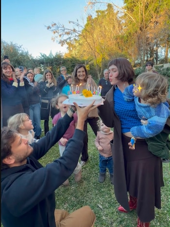 Una mujer con un bebé en brazos sopla las velas de una torta de cumpleaños, sostenida por un hombre. Varias personas, adultos y niños, los rodean en un jardín