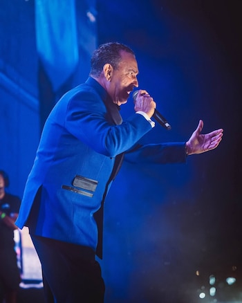 Un hombre con saco azul canta mientras sostiene un micrófono ante una iluminación de tono azul