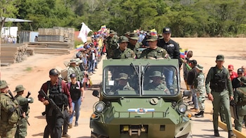 Vehículo militar blindado verde con militares adentro y alrededor, escoltado por infantería armada, entre una multitud con banderas y al lado de montones de madera