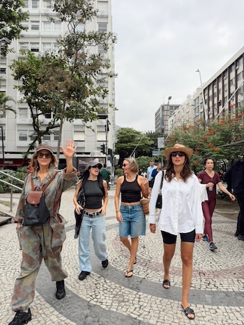 Cuatro mujeres caminan por una calle empedrada entre edificios urbanos, una de ellas saluda a la cámara levantando la mano