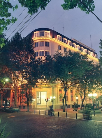 Edificio iluminado de varios pisos junto a árboles, farolas encendidas y una calle pavimentada al anochecer
