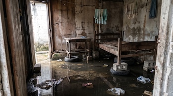 Habitación con el suelo cubierto de agua turbia, paredes con humedad y una cama y una mesa elevadas sobre bloques de hormigón y neumáticos.