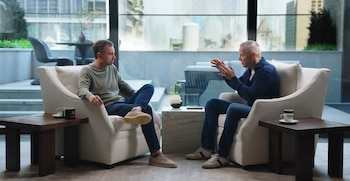 Dos hombres sentados en sillones crema conversan en un salón luminoso. Gustav Söderström, a la derecha, gesticula mientras habla. Ventanas urbanas al fondo