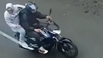 Dos personas en una motocicleta. El conductor usa casco oscuro. El pasajero viste una sudadera con capucha clara y sostiene un arma de fuego