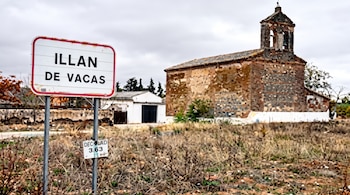 Señal de carretera blanca con marco rojo que indica 'ILLAN DE VACAS'. Detrás, una iglesia de ladrillo antigua con campanario y un terreno con hierba seca bajo un cielo nublado.