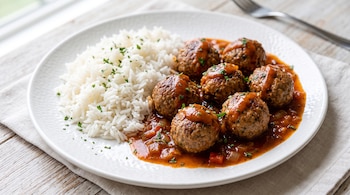 Albóndigas cubiertas con salsa de tomate junto a una ración de arroz blanco en un plato redondo sobre una mesa de madera.