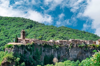 Casas de piedra y un campanario situados al borde de un acantilado basáltico, con una montaña cubierta de árboles al fondo