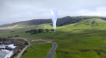 Un globo blanco transparente en el aire sobre colinas verdes, una carretera con vehículos y la costa del océano