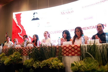 Ocho personas sentadas en una mesa con mantel rojo y blanco, botellas y plantas. Fondo con pantalla que muestra "Gobierno de México" y "Transformación Digital"