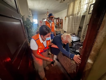 Dos personas con chalecos anaranjados y mascarillas ayudan a caminar a un hombre de edad avanzada en el interior de una vivienda