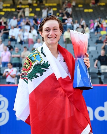 El tenista Ignacio Buse sonríe, envuelto en la bandera de Perú y sosteniendo un trofeo rojo y azul. Detrás, se ven las gradas de un estadio con público