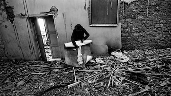 Persona con sombrero enrolla un gran retrato de un hombre en traje entre escombros de un edificio destruido. Se ven paredes derruidas y una puerta abierta