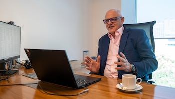 Un hombre con gafas y traje gesticula frente a una computadora portátil en un escritorio con un monitor y una taza de café