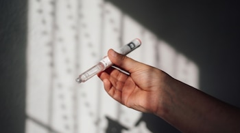 Primer plano de una mano sosteniendo una pluma de inyección médica de color blanco y transparente. El fondo es una pared clara con patrones de luz y sombra.