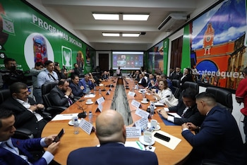 Sala de conferencias con mesa en U, múltiples personas sentadas y de pie, pantalla grande con videoconferencia, banners verdes en paredes, botellas de agua y micrófonos