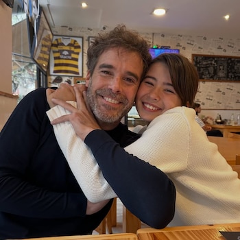 Nicolás Cabré y su hija Rufina sonríen mientras se abrazan tiernamente, sentados en una mesa de madera dentro de un local con decoración de periódicos
