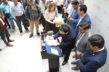 Un hombre de traje oscuro firma documentos en un atril con micrófono, mientras Greicy De Leon y varias personas lo observan en un evento legislativo
