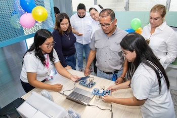 Grupo de estudiantes y adultos posan en un aula moderna con estantes llenos de kits de robótica. Sobre una mesa, un modelo de grúa y componentes tecnológicos