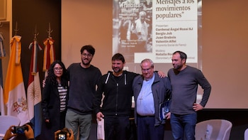 Cinco personas permanecen de pie en un escenario frente a una pantalla de proyección y dos banderas