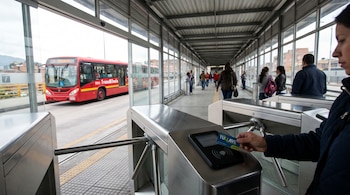 Primer plano de una persona validando una tarjeta TULLAVE en un torniquete metálico en una estación de TransMilenio, con un bus rojo visible afuera.
