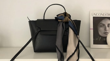 Bolso de mano negro de piel con asa superior y una solapa, decorado con un pañuelo de seda multicolor anudado, y una revista a la derecha.