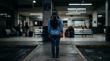 Vista posterior de una mujer con cabello largo negro, camisa y pantalón de mezclilla, de pie en la plataforma de una estación de autobuses.