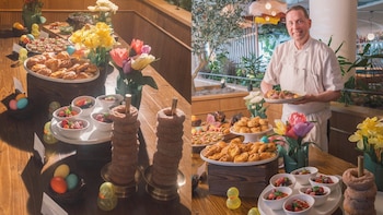 Un chef sonriente con uniforme blanco sostiene un plato de comida en un buffet festivo de Pascua con pasteles, frutas, rosquillas, huevos de colores y flores