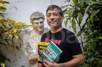 Hombre con gafas y barba sonríe y sostiene dos libros frente a una pared blanca con una escultura de busto y plantas verdes y amarillas